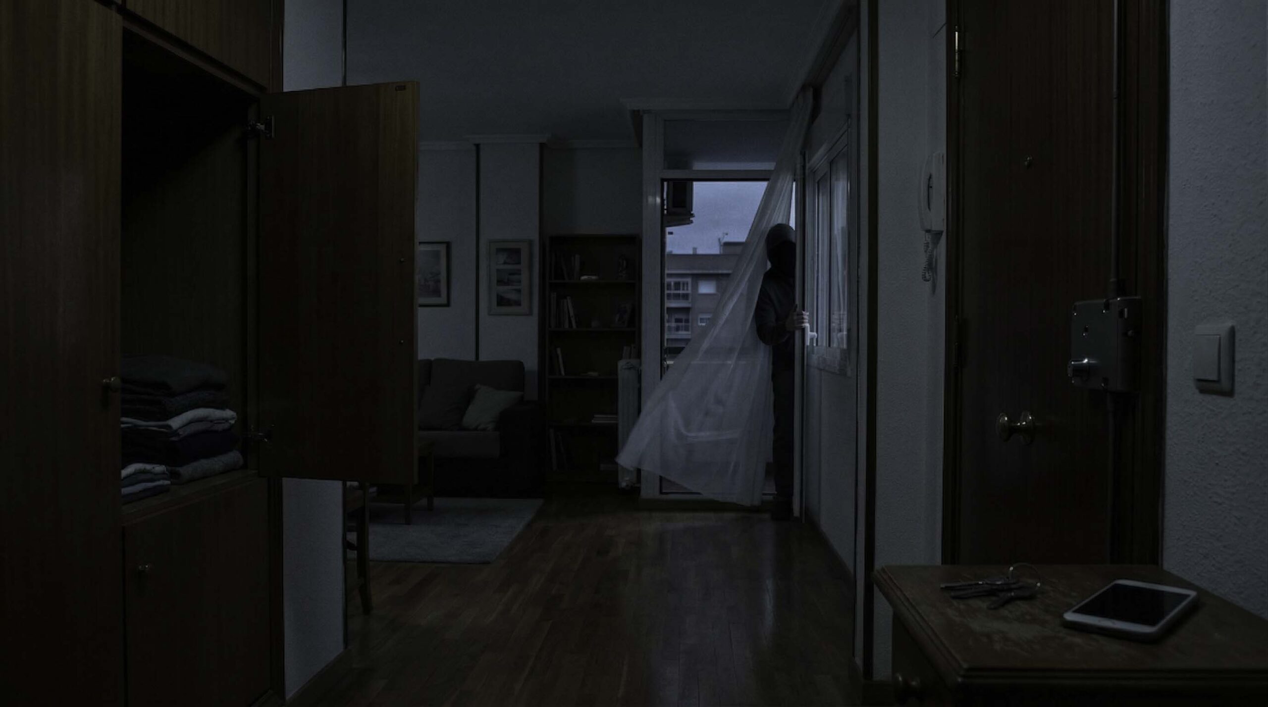 La geometría del miedo: Parte II 1 Interior de vivienda con una figura humana tras una cortina junto a la ventana, escena de suspense psicológico que sugiere intrusión confirmada en el hogar.