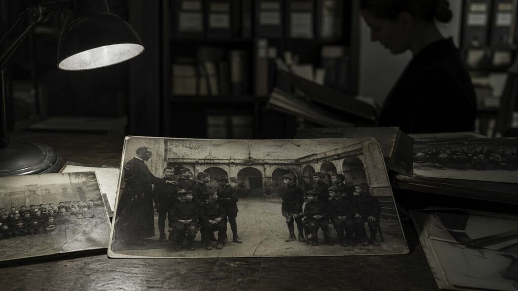 Archivo fotográfico de un internado antiguo con fotografías escolares sobre una mesa; en la imagen central, un grupo de niños deja un hueco inexplicable mientras una mujer revisa el material al fondo.
