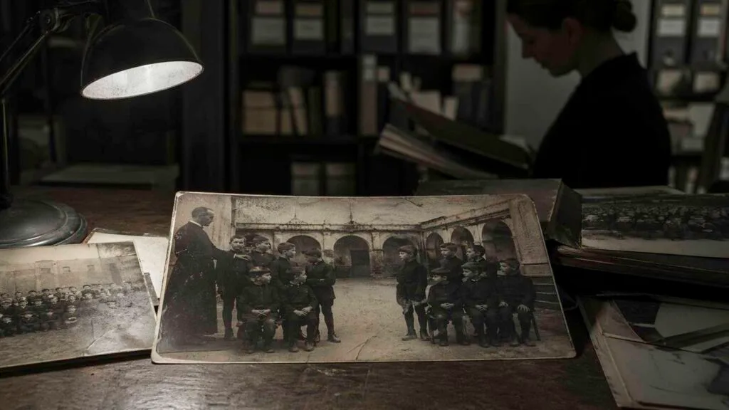 Archivo fotográfico de un internado antiguo con fotografías escolares sobre una mesa; en la imagen central, un grupo de niños deja un hueco inexplicable mientras una mujer revisa el material al fondo.