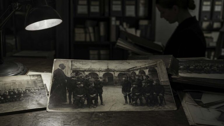 Archivo fotográfico de un internado antiguo con fotografías escolares sobre una mesa; en la imagen central, un grupo de niños deja un hueco inexplicable mientras una mujer revisa el material al fondo.