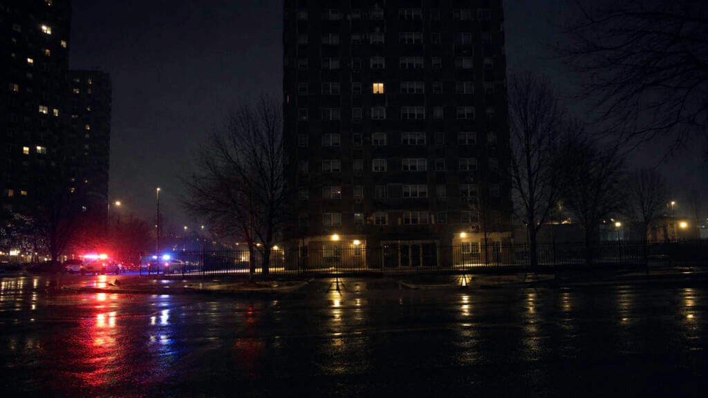 Edificio residencial de Chicago de noche, con luces de emergencia reflejadas en la calle y una ventana iluminada en la fachada.
