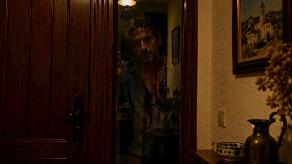 Hombre con la camisa ensangrentada pegado a la franja de cristal de una puerta de madera, de noche, mezclado con el reflejo del recibidor.