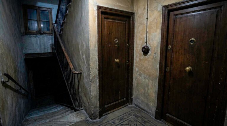 Rellano oscuro de un bloque antiguo con dos puertas de madera y una escalera que baja a la penumbra